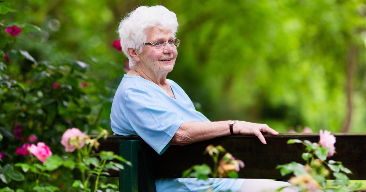 A very cute senior woman with short grey hair sits on a black bench surrounded by greenery and flowers, gazing thoughtfully in a specific direction.