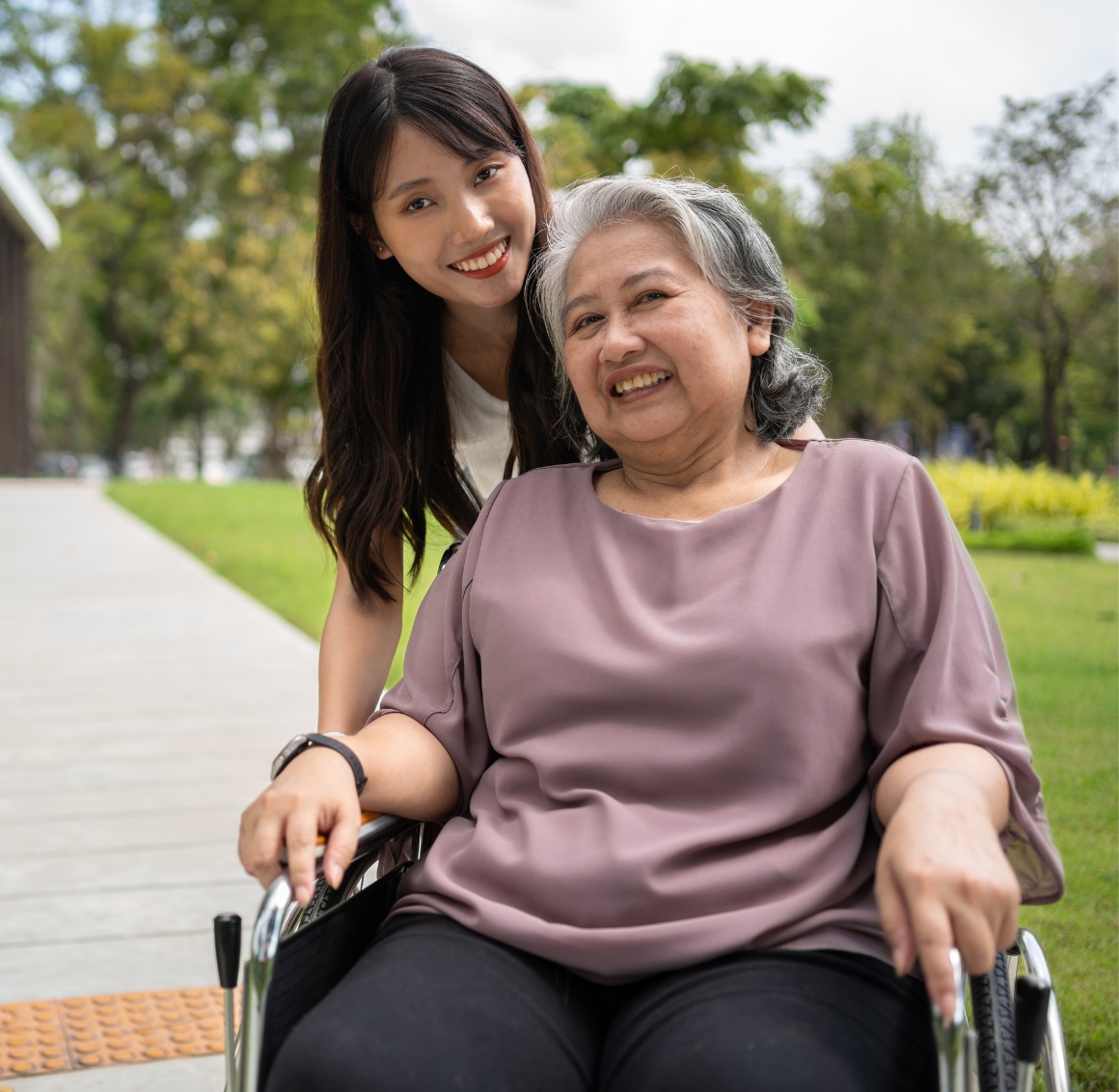 An Asian young woman with black hair leans over a senior woman with short grey hair seated in a wheelchair outdoors, surrounded by greenery. They share a quiet, tender moment in the natural setting.