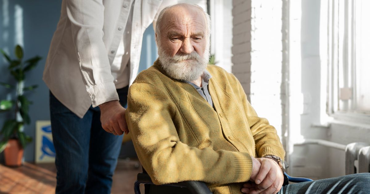 A man stands behind a senior man with dementia who is seated in a wheelchair indoors, wearing a yellow cardigan. The atmosphere is calm and supportive.
