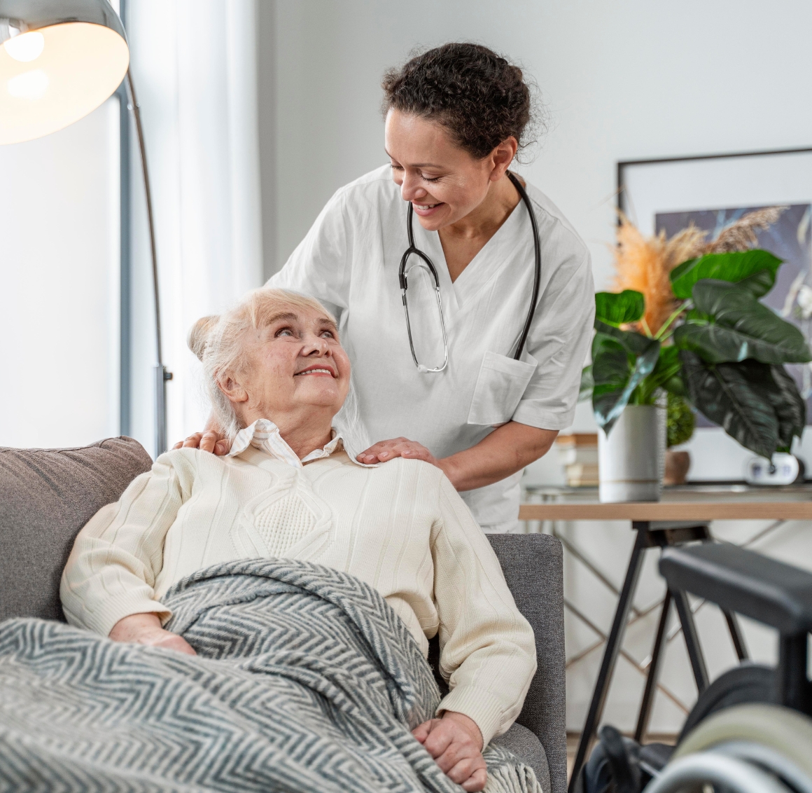 A caregiver in white scrubs stands behind an elderly woman seated on a couch in a cozy living room, gently resting her hands on the woman’s shoulders while looking over her with care.