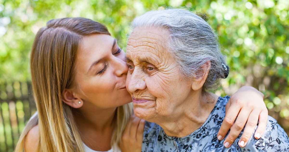 A young woman lovingly kisses the cheek of a smiling senior woman outdoors in a green, leafy setting, with her arm wrapped gently around her shoulders.