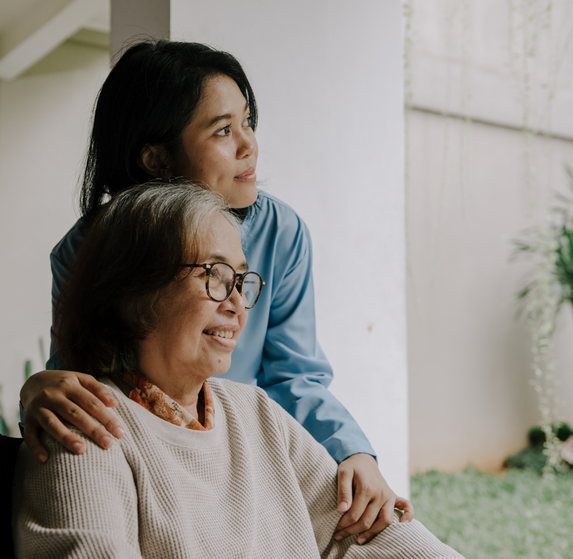 An Asian caregiver with black hair stands behind a senior Asian woman seated in a wheelchair outside on a driveway. They are both outdoors, with the caregiver positioned attentively behind the wheelchair.