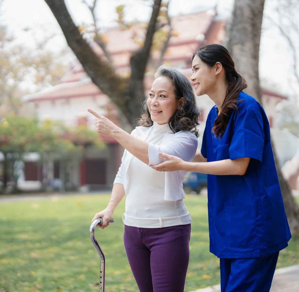 An Asian caregiver with black tied hair wearing blue scrubs stands next to an elderly Asian woman as they walk together outside; the senior points toward something in the neighborhood, capturing a moment of shared curiosity and connection.