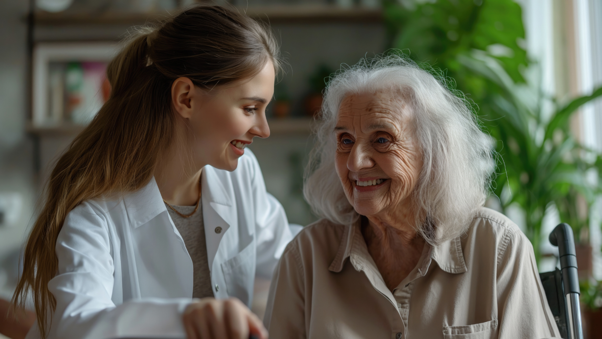 A young caregiver with long black hair sits next to an elderly woman with grey hair on a couch indoors, both looking at each other with warmth and love, reflecting a strong bond of care and connection.
