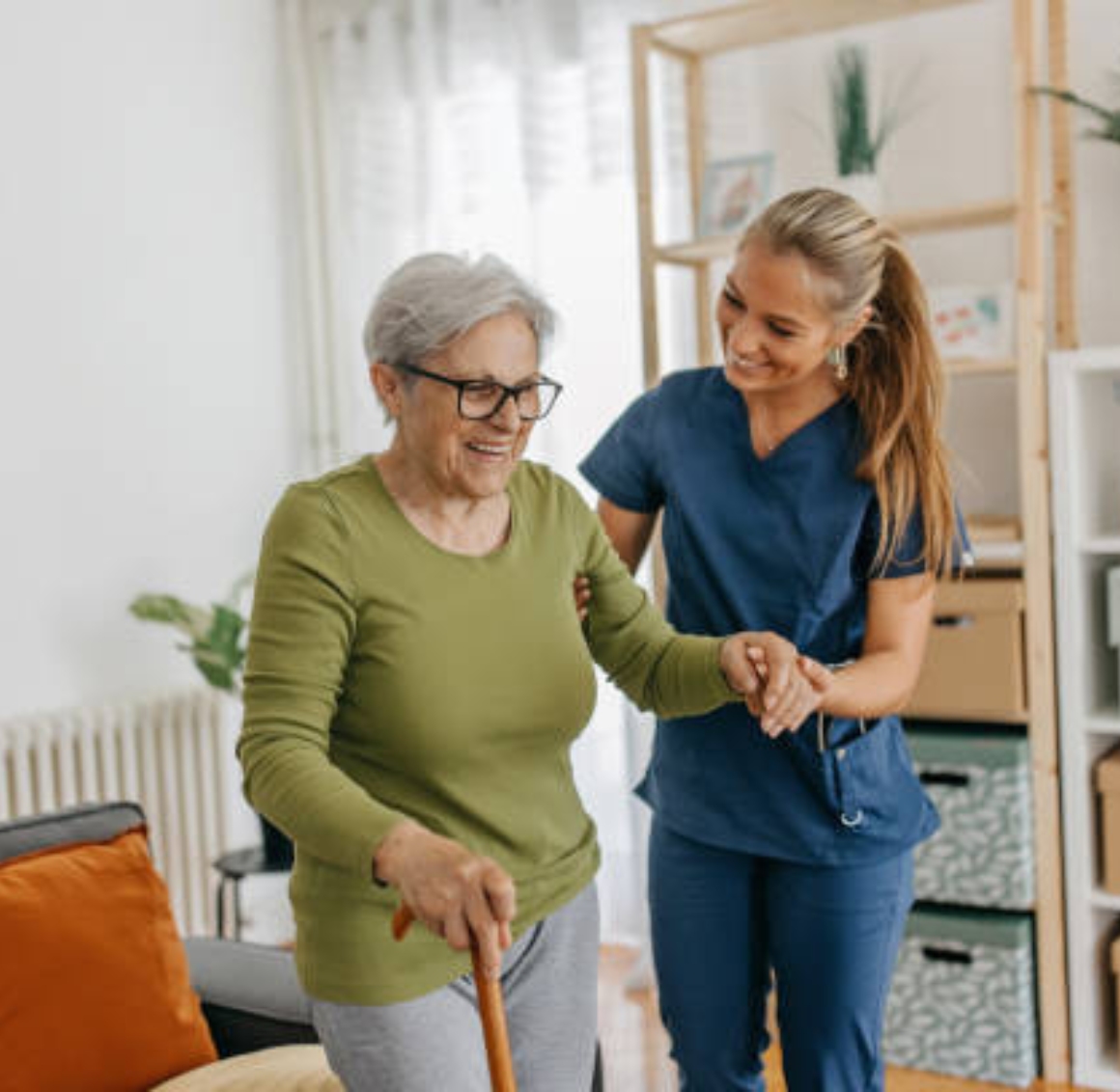 A young female caregiver in dark blue scrubs helps an Asian senior woman stand with support indoors, holding her hands gently to provide balance and encouragement.