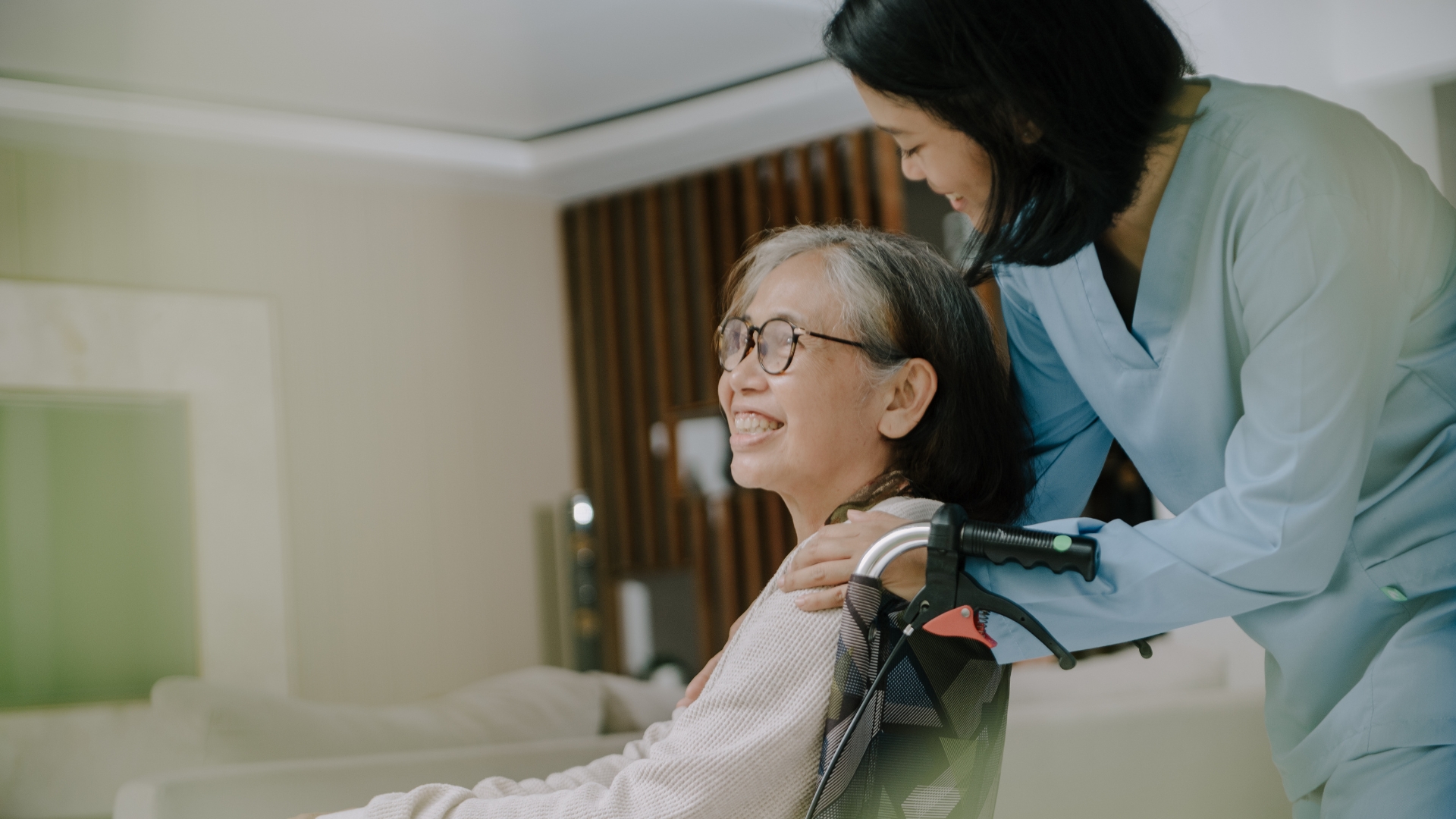 An Asian caregiver stands behind a senior Asian woman in a wheelchair as they exchange a warm, caring glance in a comfortable living room. This image captures the essence of senior home care in Gretna, highlighting trust, connection, and compassionate support in a familiar home setting.