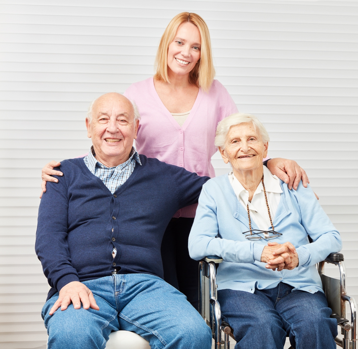 A caregiver poses with a seated senior couple, smiling warmly. The bright, welcoming scene highlights compassionate senior home care in Carlisle.
