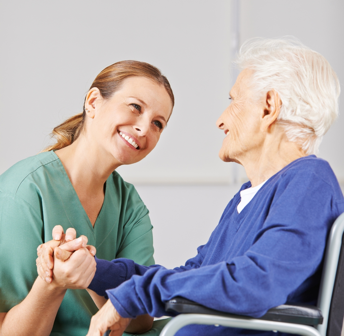 A caregiver in green scrubs holding hands with an elderly woman in a wheelchair, both smiling at each other. This touching moment represents attentive and personalized home care in LaVista.