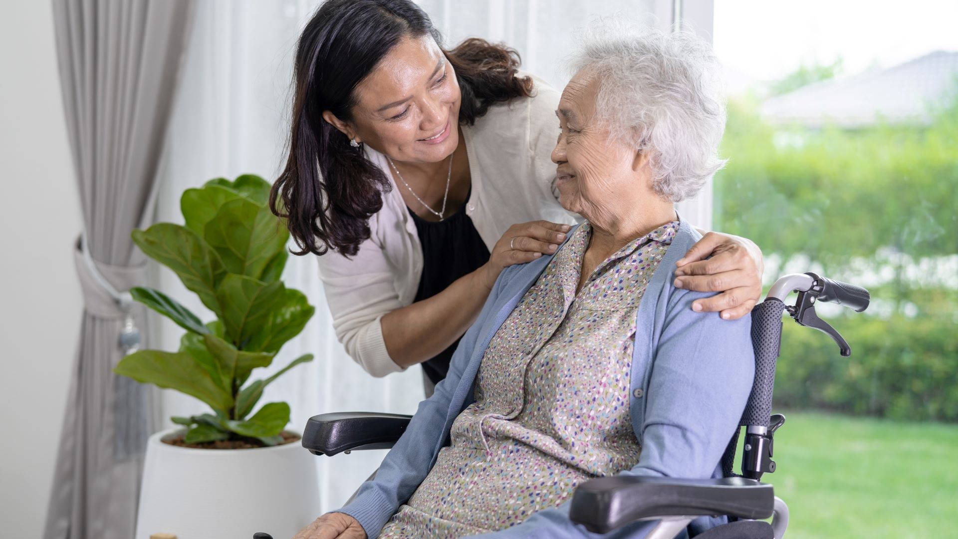 An Asian caregiver wraps her arms gently around an Asian senior woman who is seated in a wheelchair. The embrace conveys warmth, support, and a sense of close companionship.