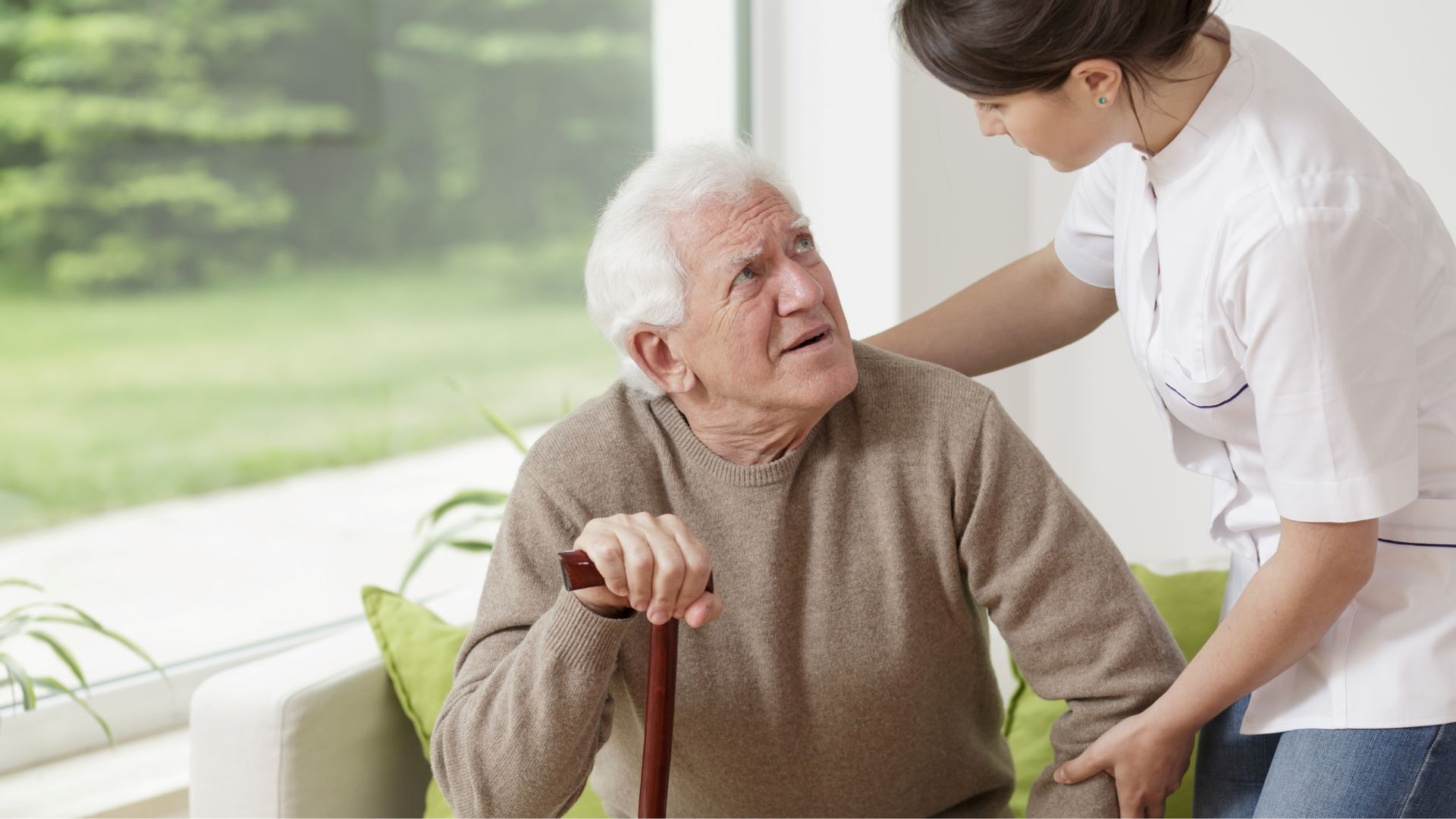 An Asian female caregiver provides supportive care to a senior man as he looks at her while sitting on an indoor sofa. The moment reflects compassion and trust, showcasing the personalized attention offered through senior home care in Waukee.