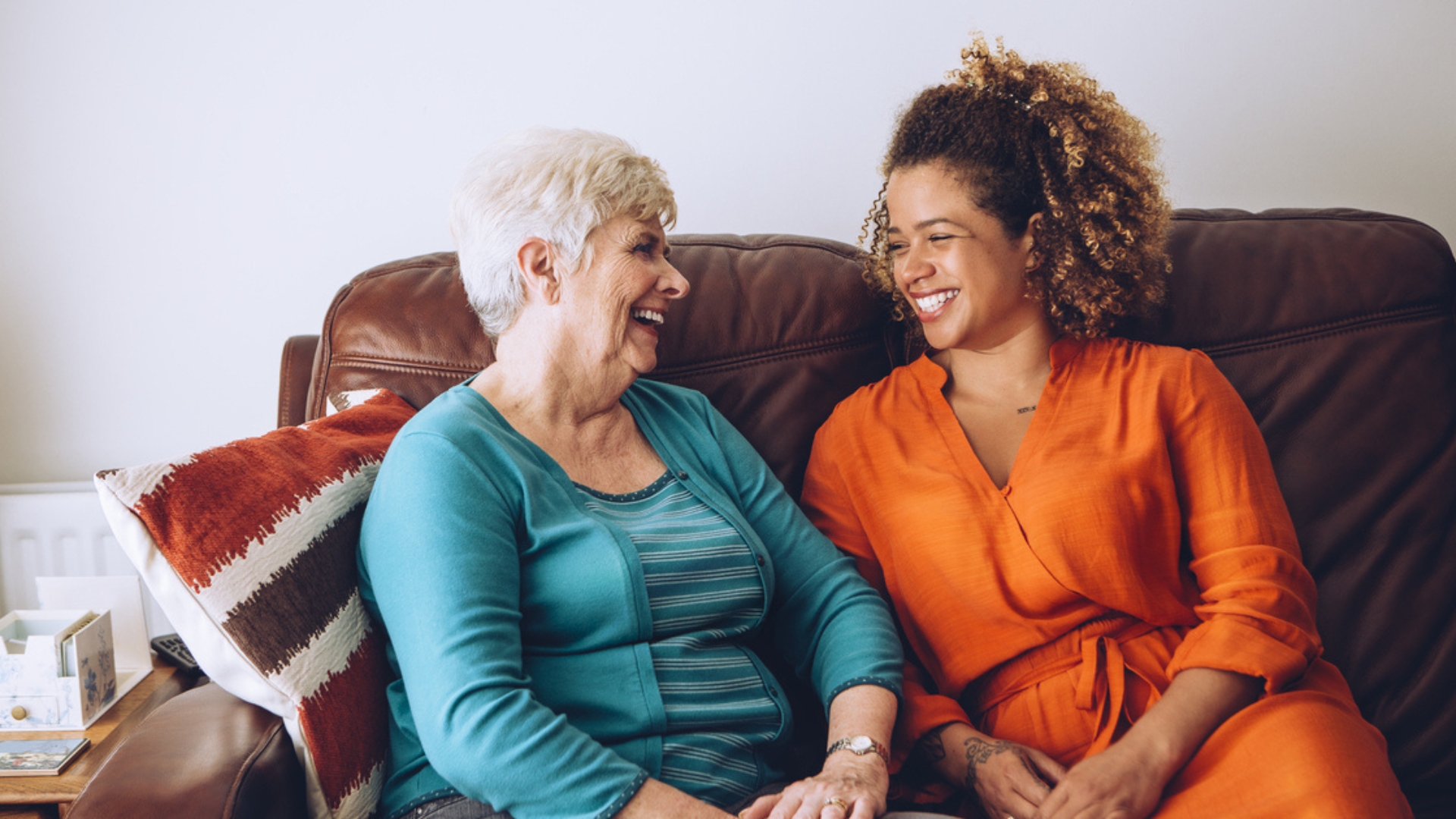 A cheerful elderly woman and a younger caregiver sit together on a brown leather couch, smiling warmly at each other. The scene reflects a supportive and friendly environment, capturing the essence of senior home care in Waverly.
