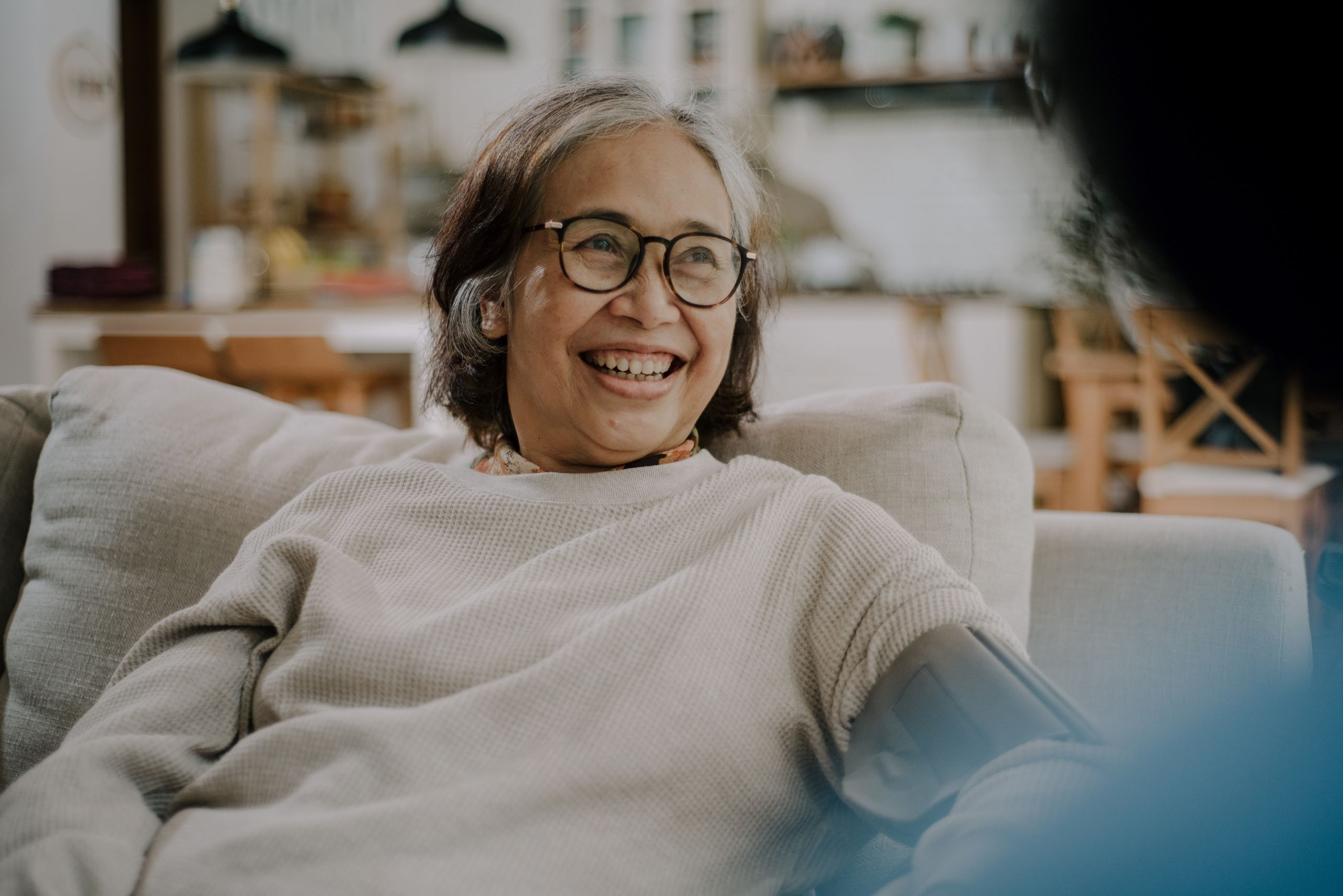 A smiling elderly woman wearing glasses sits comfortably on a couch in a cozy, well-lit home setting. Her relaxed and joyful expression reflects the positive impact of compassionate senior home care in Grimes, highlighting comfort, dignity, and personalized support in a familiar environment.