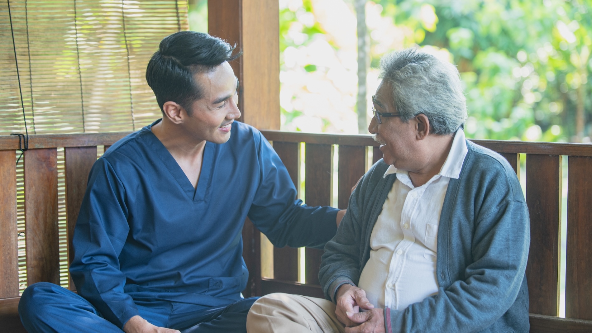 A male caregiver in navy blue scrubs sits beside an elderly man on a wooden bench, gently placing a hand on his shoulder as they share a warm and friendly conversation. The relaxed, outdoor setting surrounded by greenery reflects the compassionate and personal nature of senior home care in Norwalk, IA, emphasizing trust, connection, and supportive companionship.