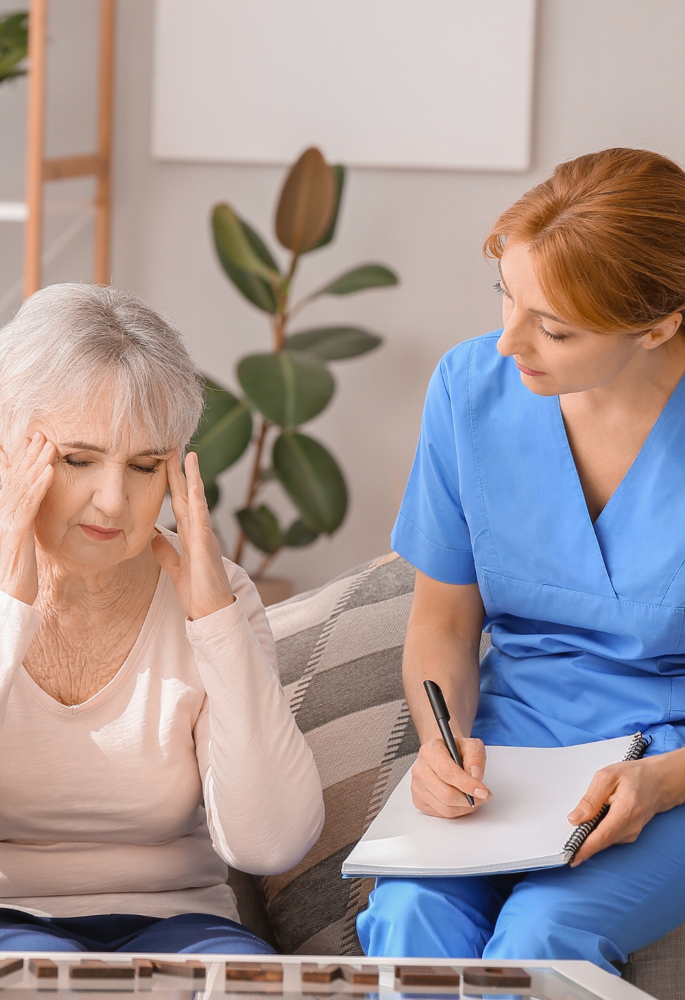 A concerned elderly woman holds her head while a caregiver in blue scrubs takes notes beside her. Dementia Care in Lincoln offers professional support to help seniors manage memory loss, confusion, and daily challenges with patience and understanding.