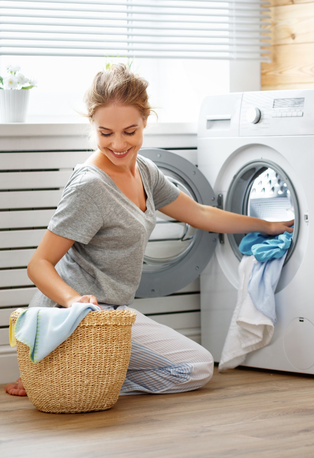A smiling woman kneels by a washing machine while placing clothes inside a laundry basket. Homemaker Services in Lincoln provide reliable help with laundry and household chores so seniors can enjoy a clean and organized home.