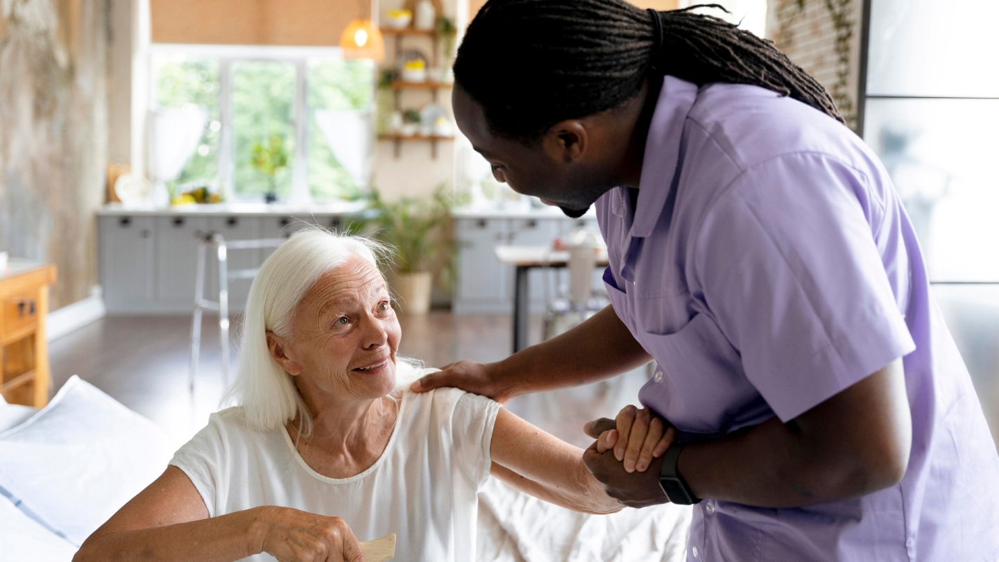 A professional caregiver offering support to an elderly woman at home, both smiling warmly at each other. The setting is a bright, cozy kitchen and living space, highlighting the compassionate and personalized approach of senior home care in Eagle.