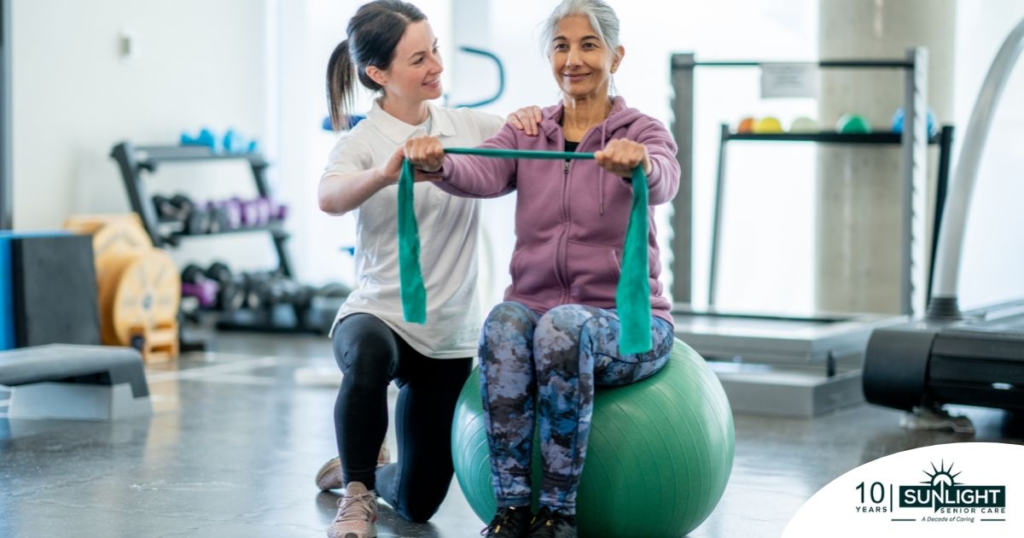 A care provider helps an older woman exercise with a resistance band and an exercise ball, representing how exercise can help with senior fall prevention.